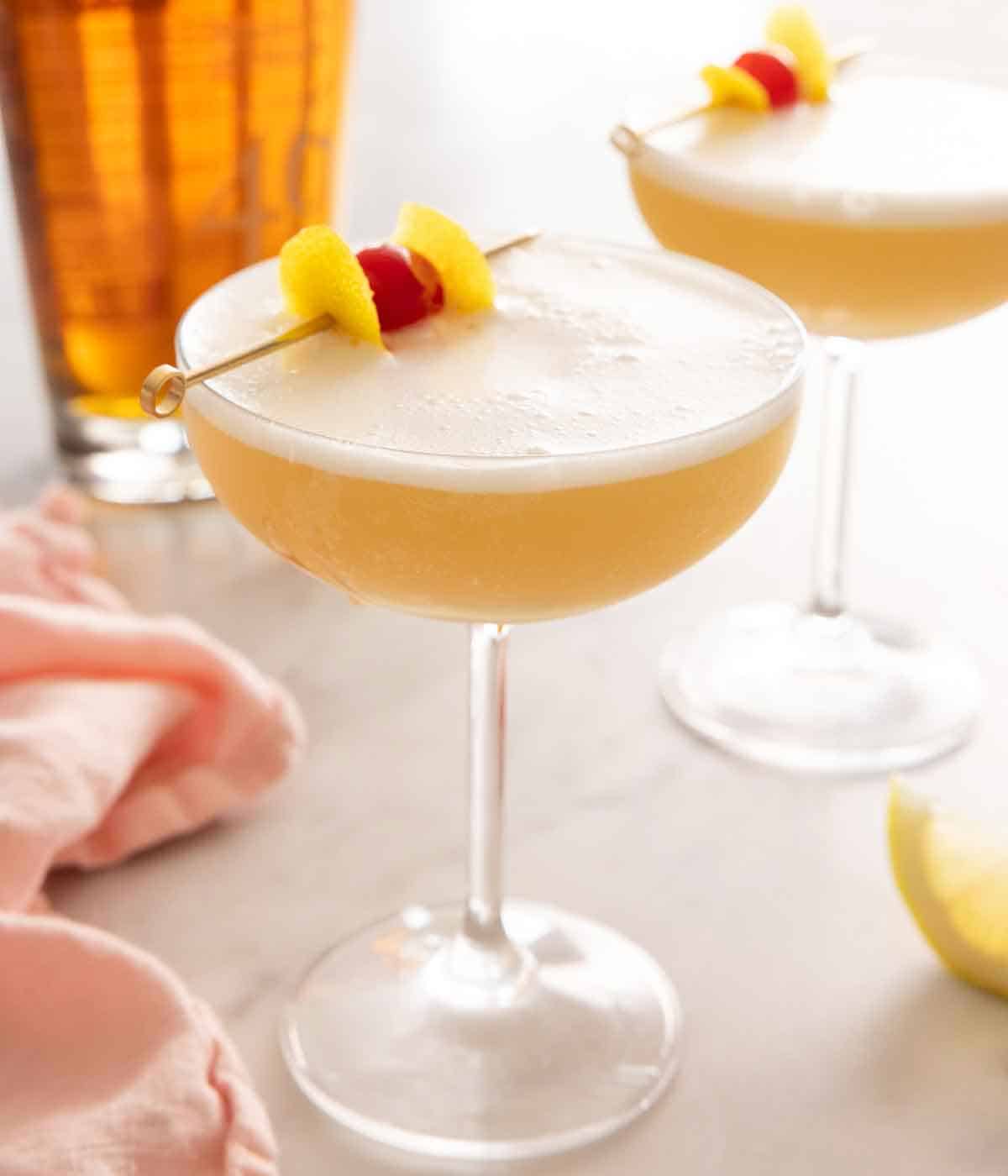 Two whiskey sours in cocktail glasses with cocktail picks with lemon peel and maraschino cherry as garnish.