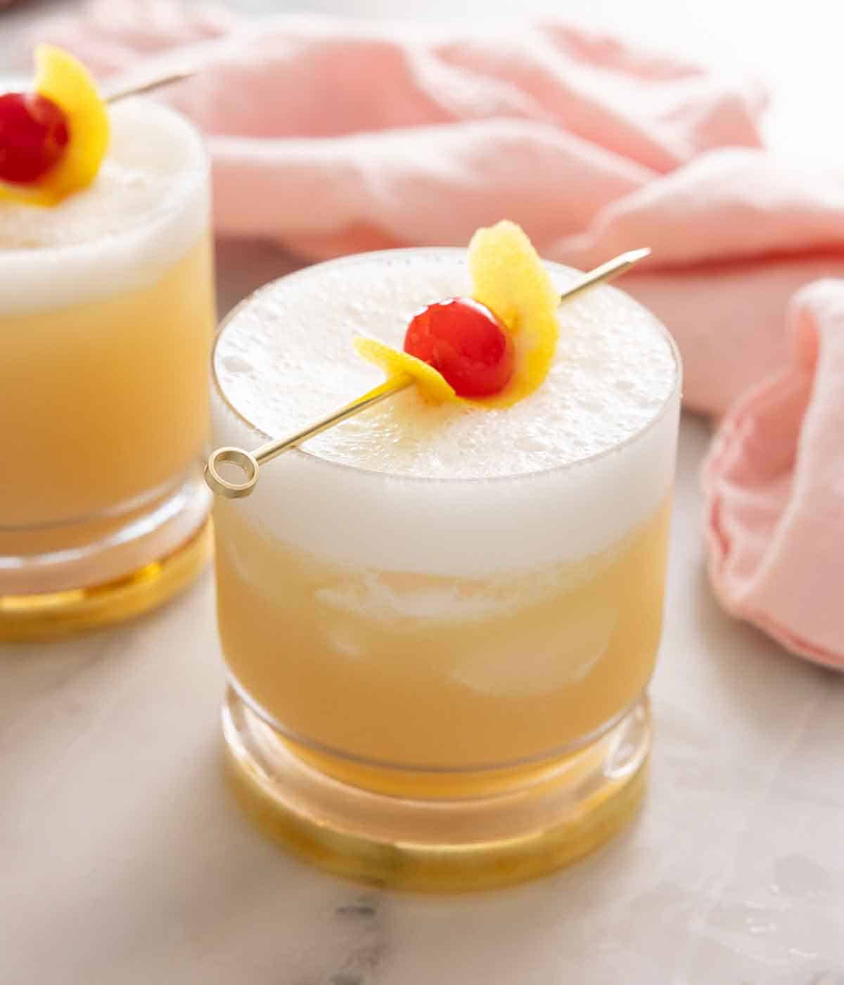 Two whiskey sours, one in front, with a lemon peel and maraschino cherry garnish beside a pink linen napkin.