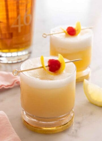 Two glasses of whiskey sour with cocktail picks with lemon peel and maraschino cherry as garnish on top.