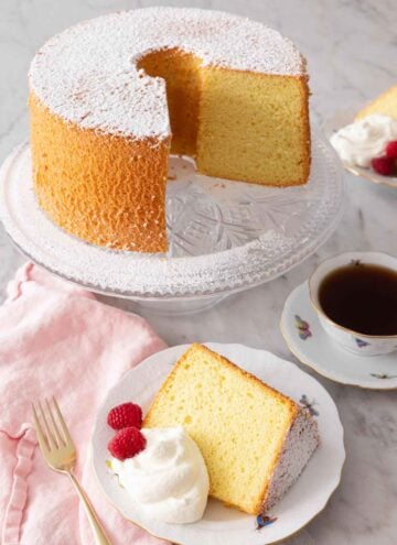 A chiffon cake on a stand, cut with a slice plated in front with whipped cream and berries.