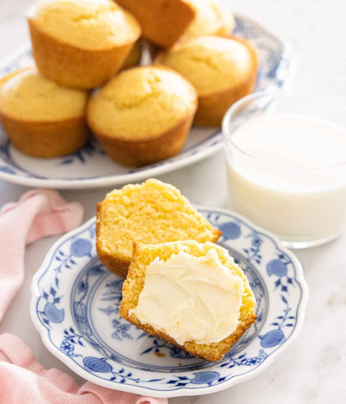 A plate with a cornbread muffin cut in half with butter spread on it. More cornbread muffins and a glass of milk in the background. 