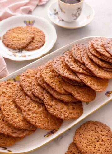 A platter with multiple lace cookies in front of a plate with two cookies and coffee.