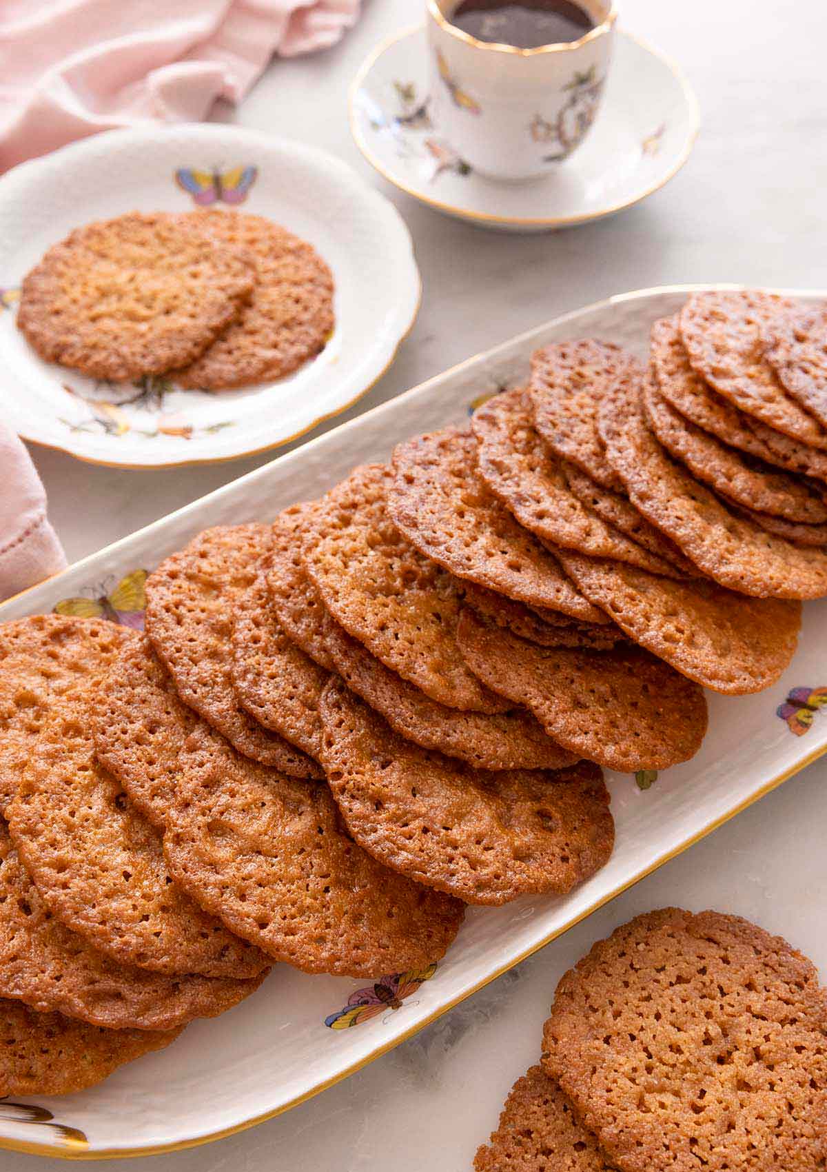 A platter with multiple lace cookies in front of a plate with two cookies and coffee.