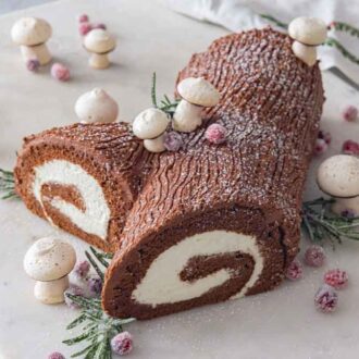A marble serving board with a Buche de Noel with meringue mushrooms, sugared cranberries, and rosemary scattered around the cake.
