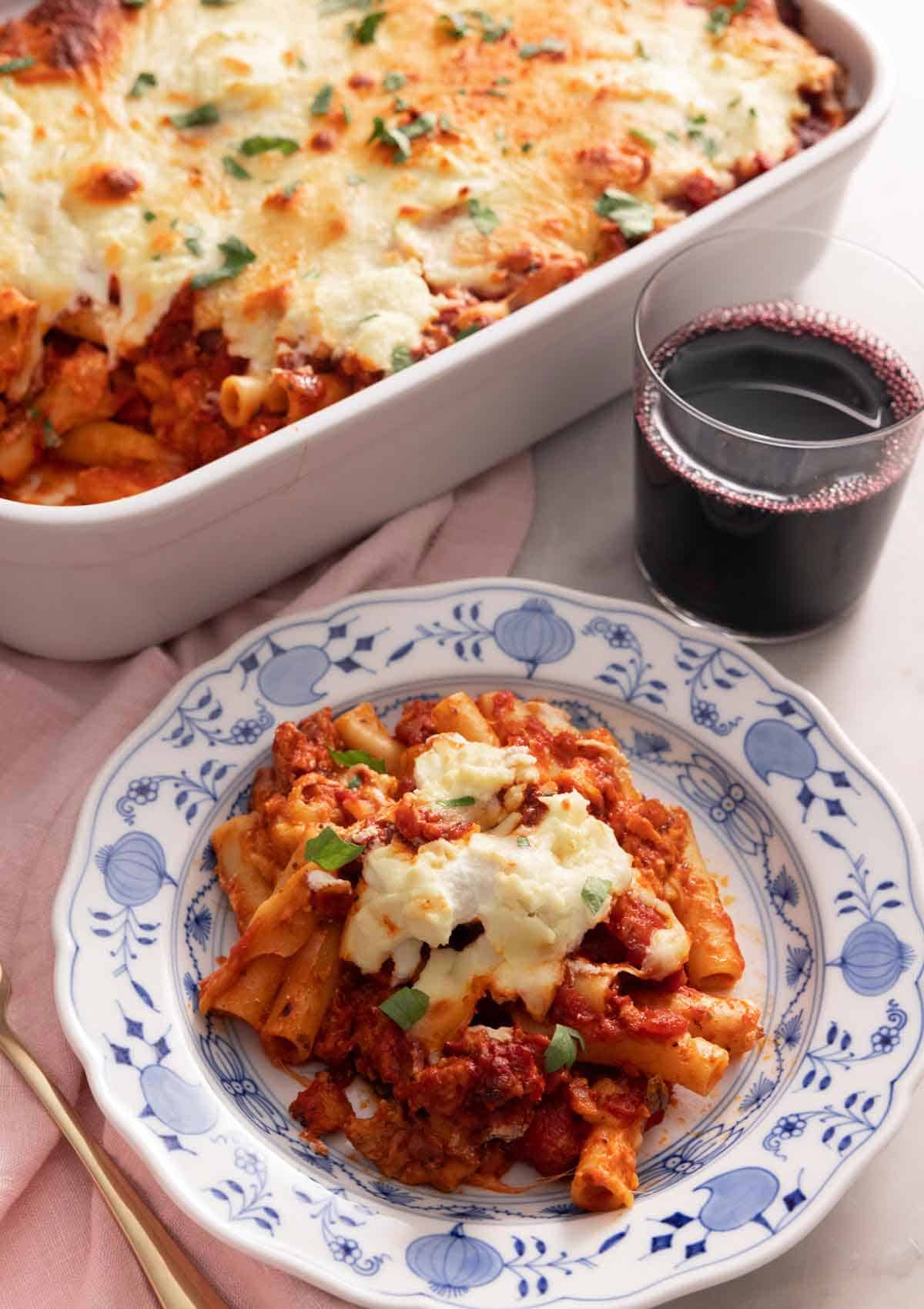 A plate with a serving of baked ziti by a glass of wine and baking dish.