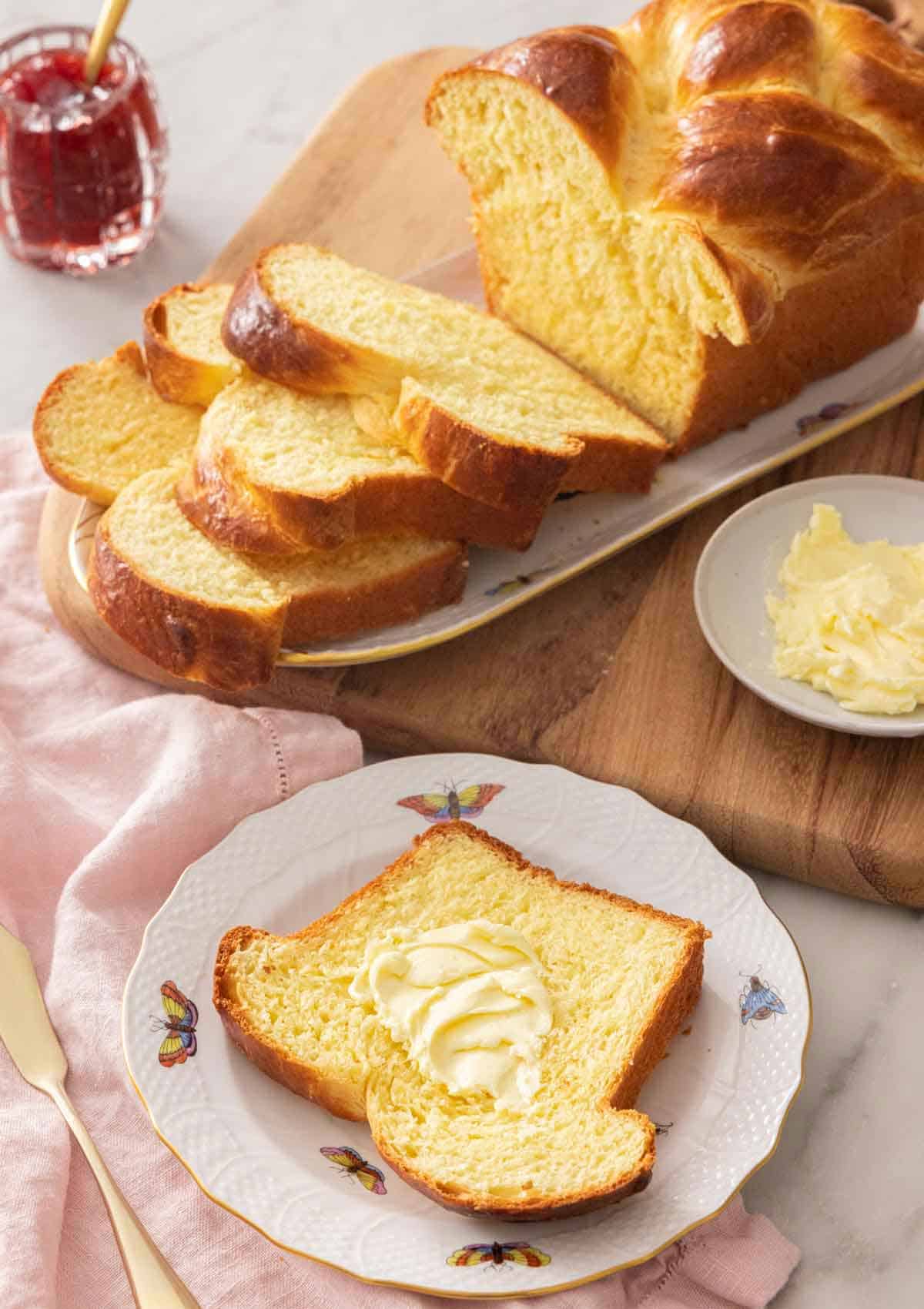A slice of brioche bread with butter on it, in front of a sliced loaf.