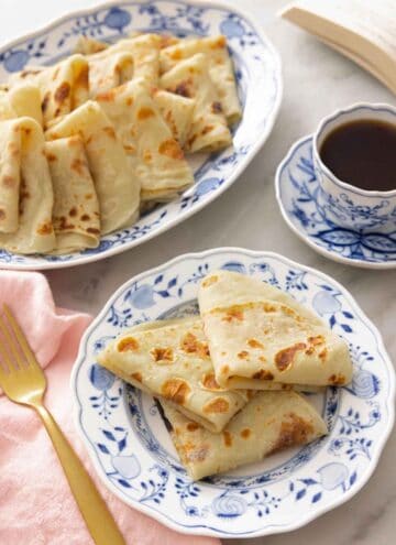 A plate with three folded lefse by a mug of coffee and a platter.