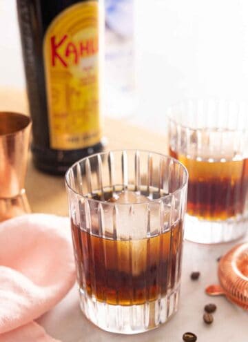 Two glasses of Black Russians with one in front. A bottle of Kahlua in the background.