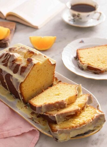A loaf of orange cake with three slices cut by a mug of coffee and orange wedges.