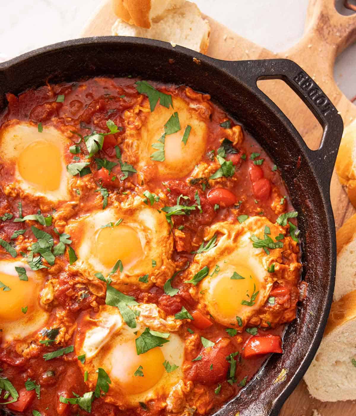 Close up of a pan of shakshuka with parsley garnish on top, on a serving board with slices of bread.