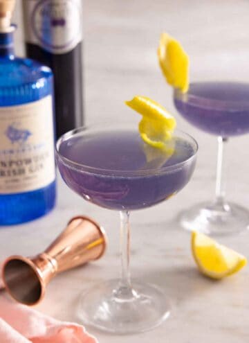 Two glasses of Aviation cocktail with lemon twist garnish by the bottles of liqueur in the back.