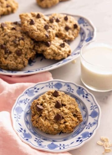 A plate with a banana oatmeal cookie by a glass of milk and a platter with more.
