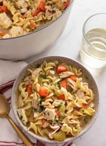 A bowl of chicken noodle soup by a glass of wine.