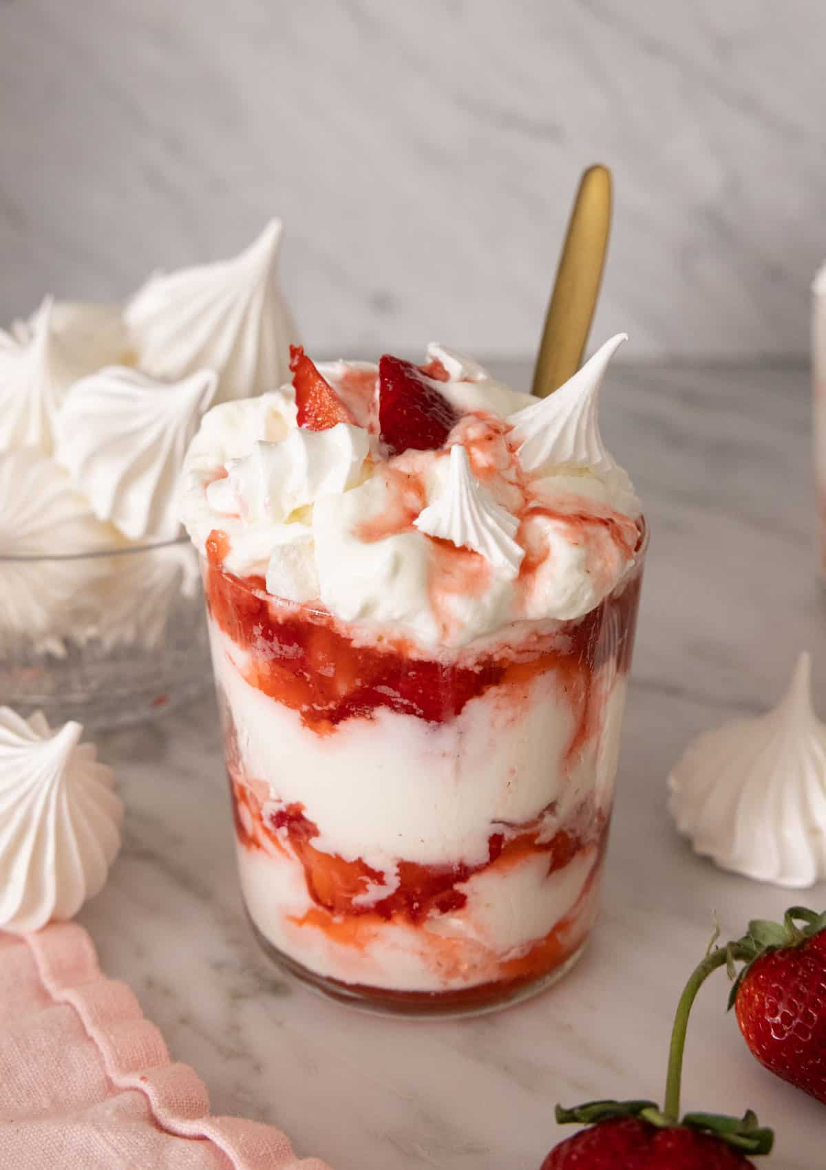 A glass of Eton mess with one in front with strawberries and meringue cookies around it.