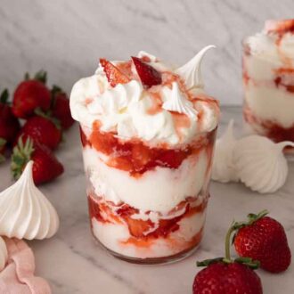 Two glasses of Eton mess with strawberries and meringue cookies scattered around.