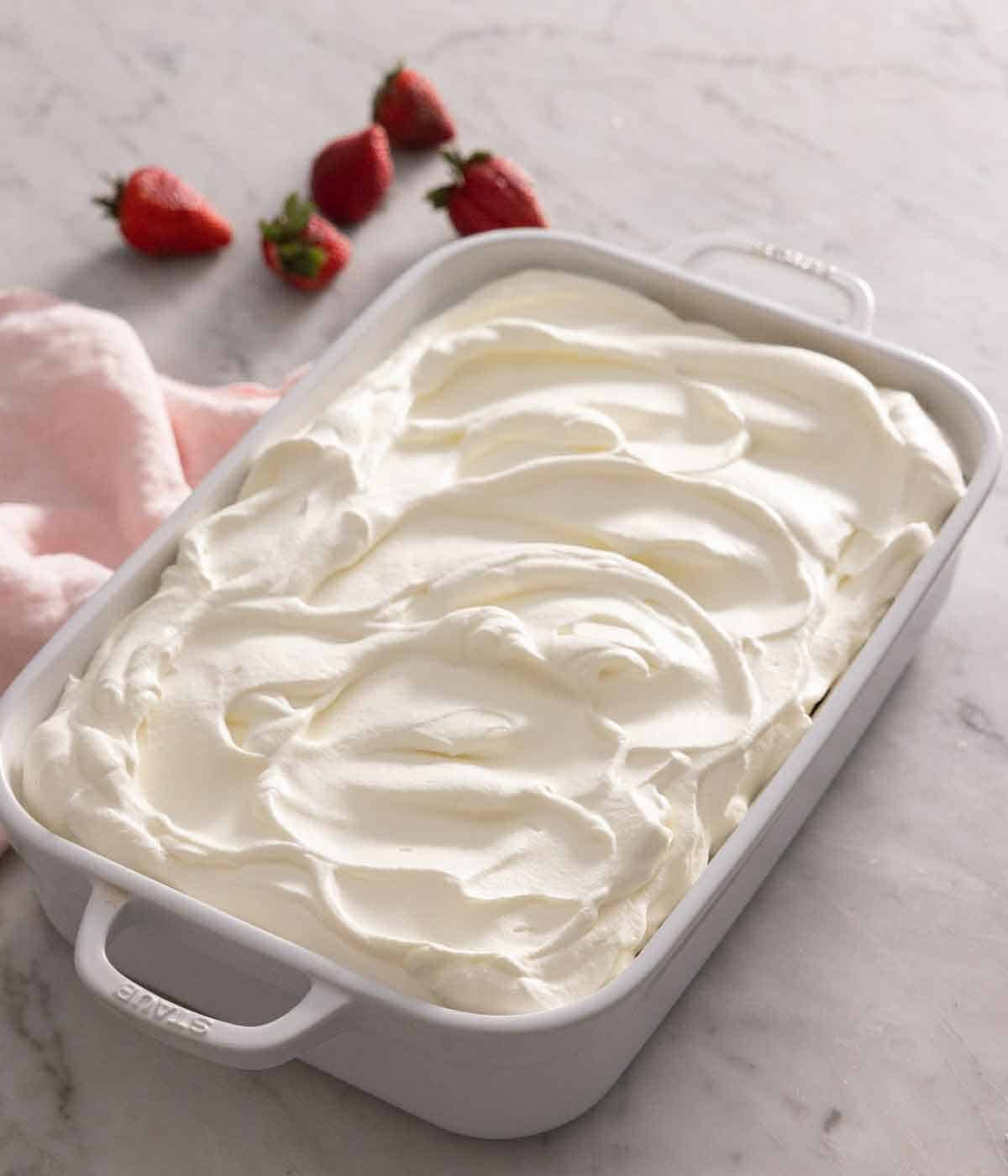 A baking dish with tres leches cake with frosting on top and strawberries in the background.