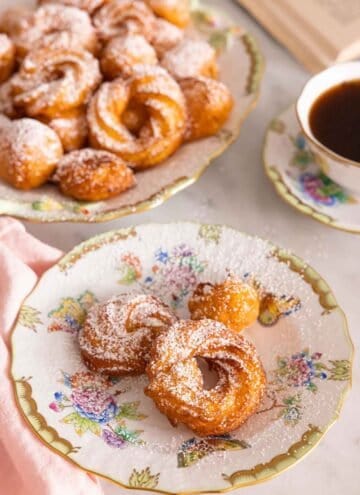 A plate of zeppole with more in the back with powdered sugar dusted on top.