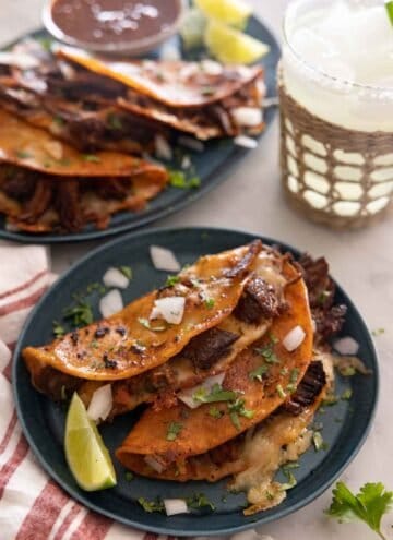 A plate with two birria tacos in front of a drink and platter of more tacos.