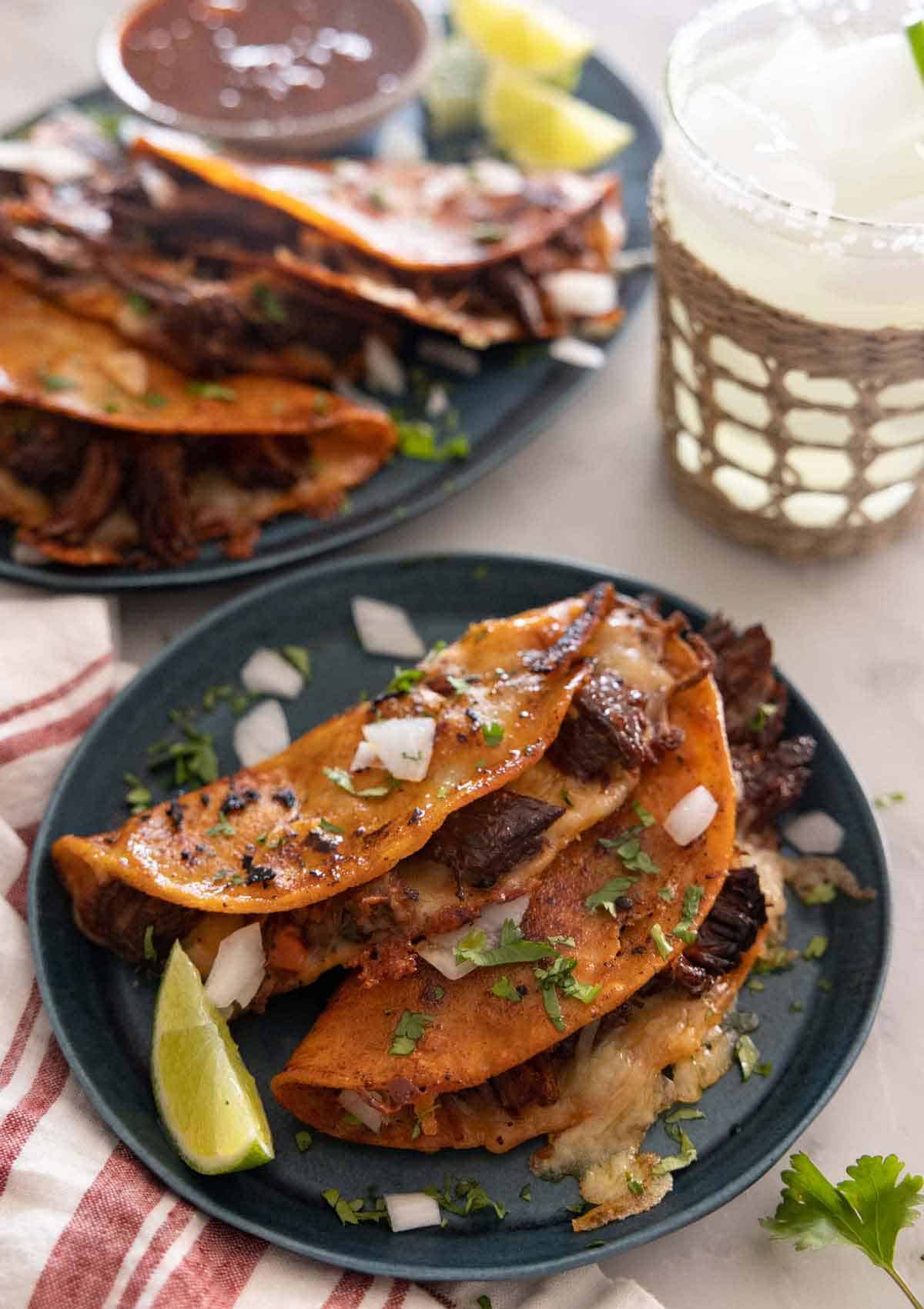 A plate with two birria tacos in front of a drink and platter of more tacos.