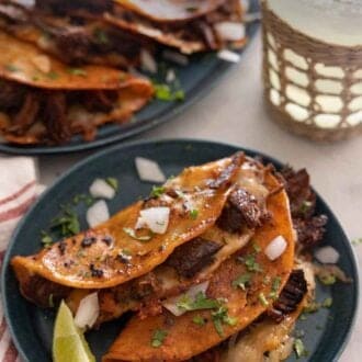Pinterest graphic of two birria tacos on a plate with a lime wedge in front of a platter with more tacos.