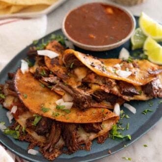 A platter of birria tacos with a bowl of dip and lime wedges.