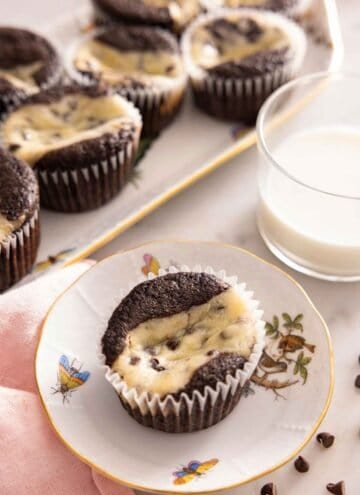 A plate with a black bottom cupcakes by a cup of milk and a platter of more cupcakes.