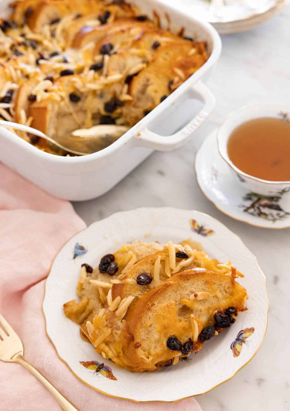 A plate with a serving of capirotada in front of a cup of tea and a baking dish.