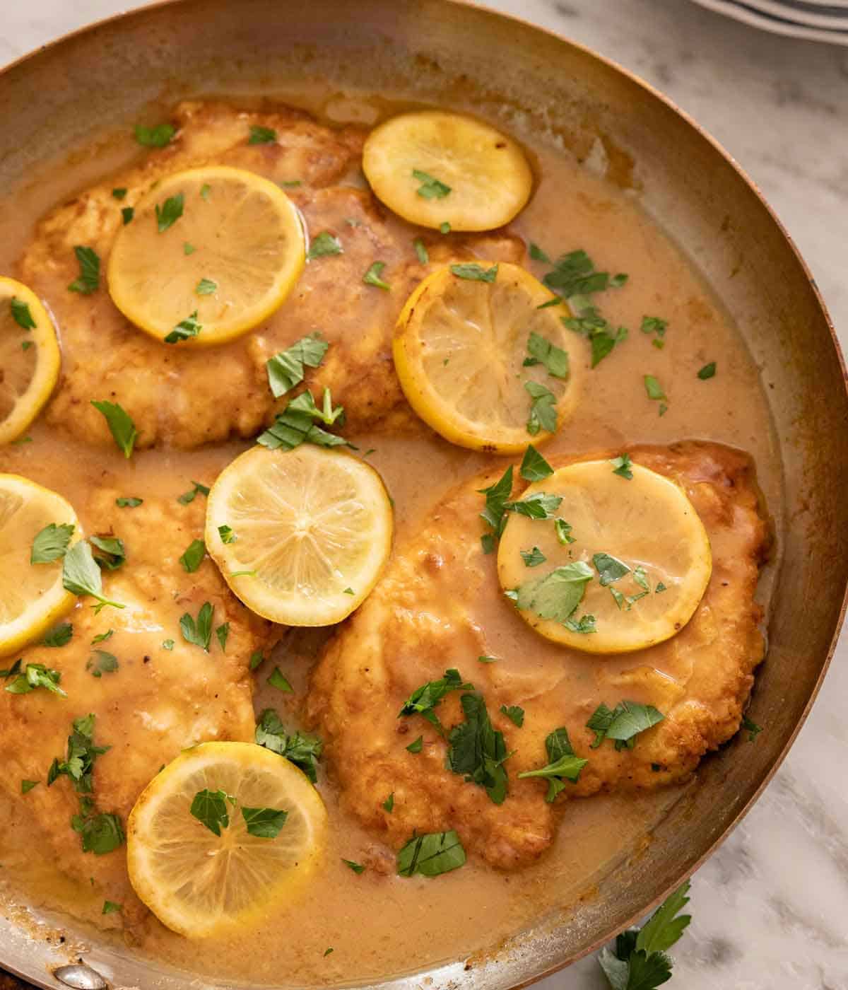 A skillet of chicken Francese with sliced lemons and parsley on top.