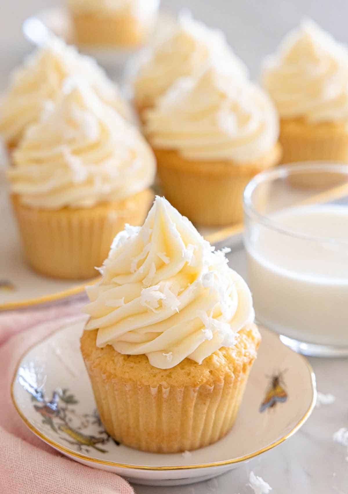 A plate with a coconut cupcake with more in the background along with milk.