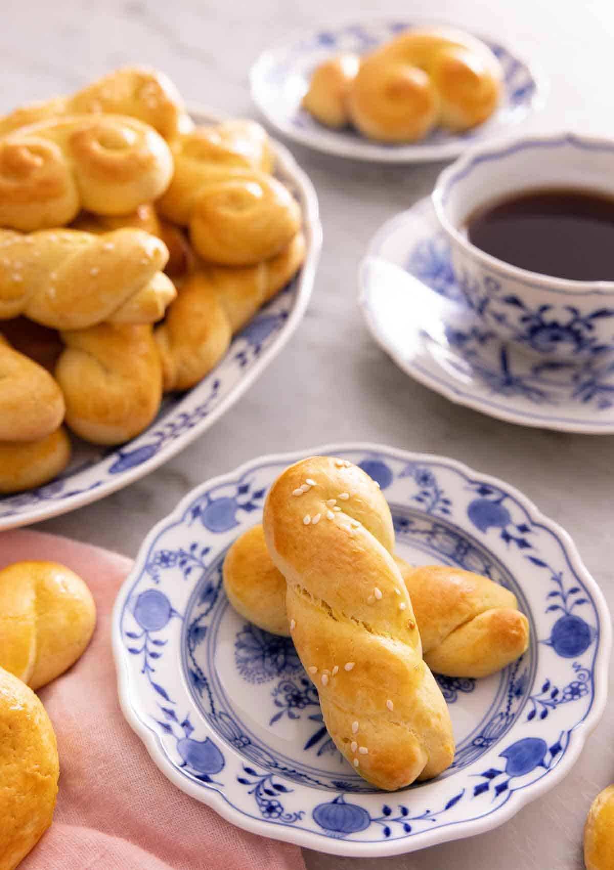A plate with two koulourakia by a mug of coffee and a platter more.