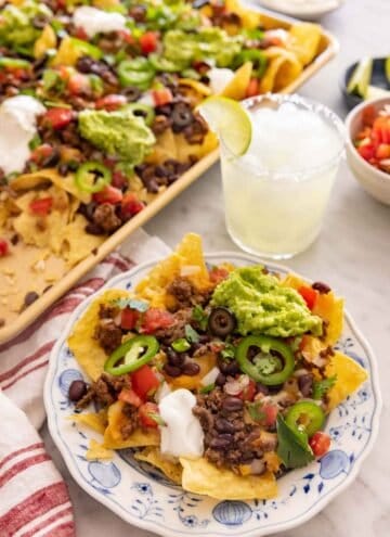 A plate with a serving of nachos by a drink and sheet pan of more nachos.