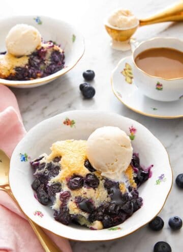 A bowl if a serving of blueberry cobbler with a scoop of vanilla ice cream on top. Mug of coffee, cobbler, and more ice cream in the background.