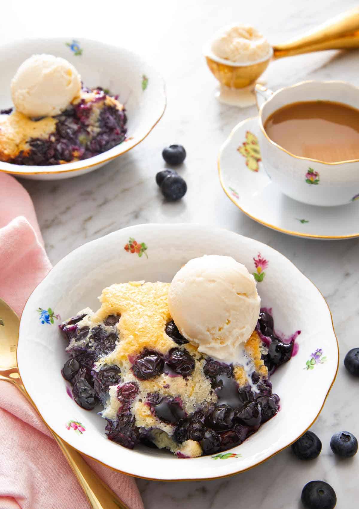 A bowl if a serving of blueberry cobbler with a scoop of vanilla ice cream on top. Mug of coffee, cobbler, and more ice cream in the background.