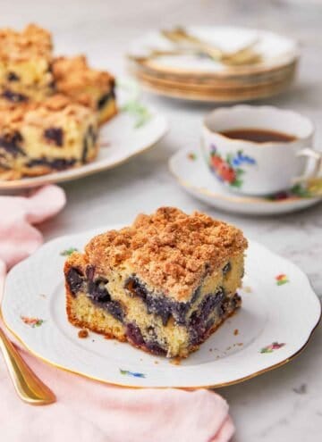 A square piece blueberry coffee cake on a plate in front of a cup of coffee.