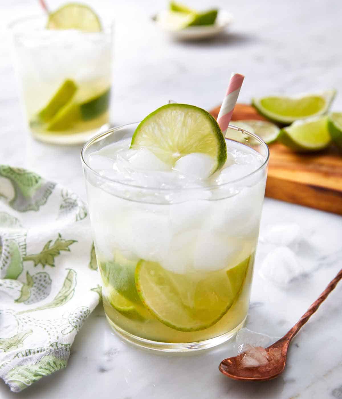 A glass of caipirinha with with a pink and white straw inserted. Fresh lime in the background on a cutting board.