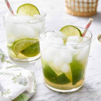 Two glasses of caipirinha with a straw in each and a small plate of cut limes in the background.
