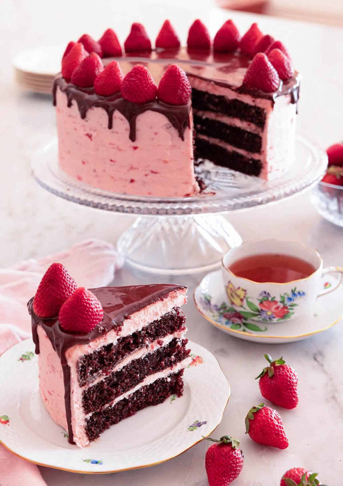 A slice of chocolate strawberry cake on a plate in front of a cake stand with the rest of the cake.