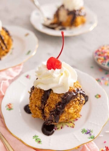 A plate with a ball of fried ice cream with chocolate drizzle and whipped cream on top with a cherry.