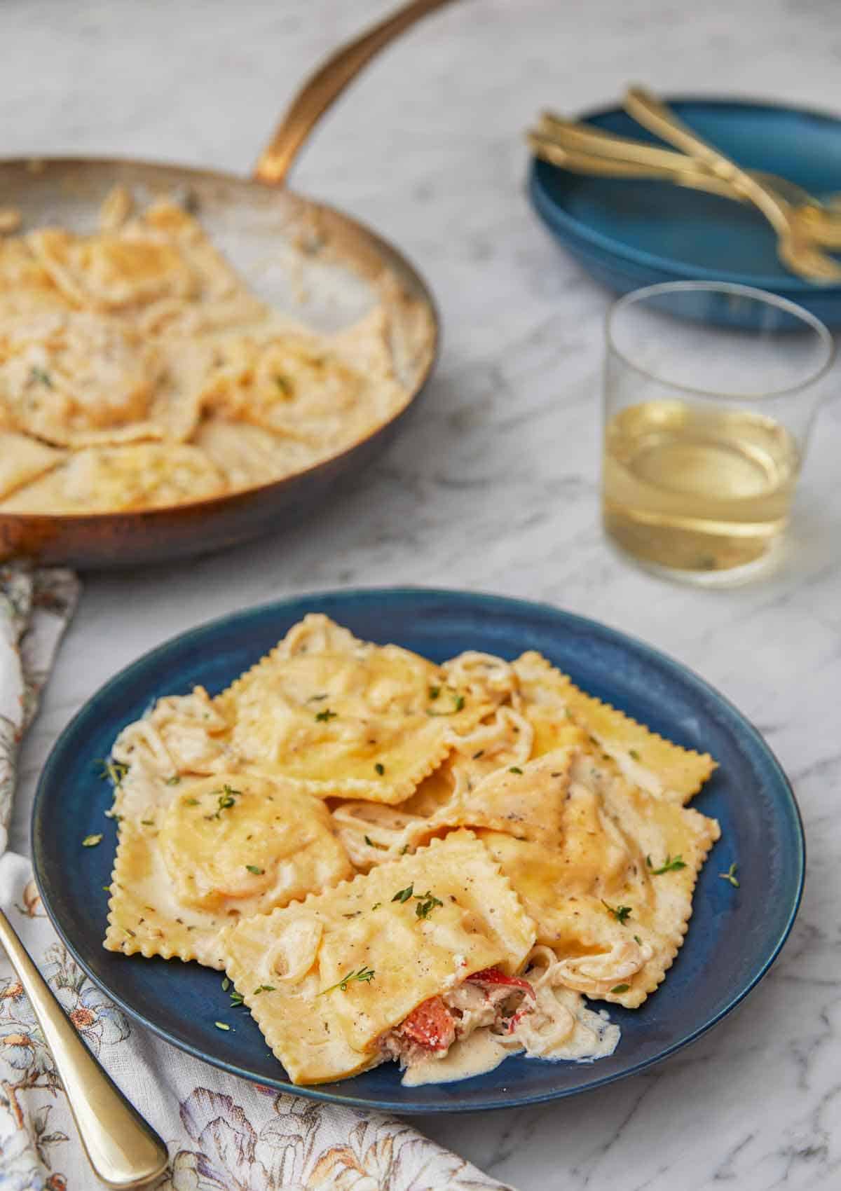 A plate of lobster ravioli by a glass of wine and a skillet with pasta.