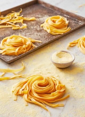 Multiple bundles of pasta dough on a sheet pan with one in the foreground with a small bowl of semolina flour.