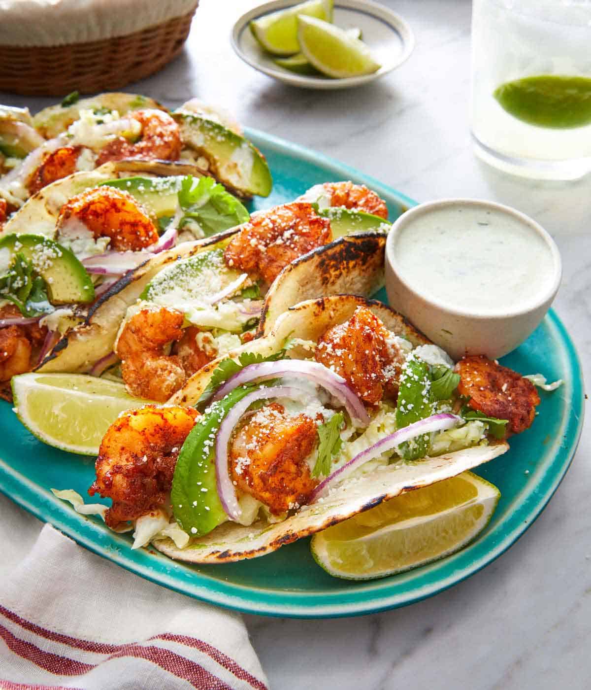 An oval platter with multiple tacos filled with grill, avocado, red onions, cabbage, and cotija cheese with a small bowl of dip on the side.