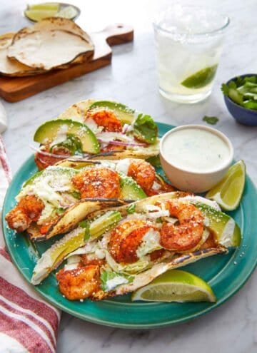 A plate with three shrimp tacos with a dip on the side with cotija cheese sprinkled on top of the tacos.