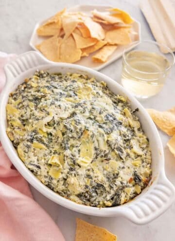 A white baking dish of spinach artichoke dip with a plate of chips and glass of wine behind it.