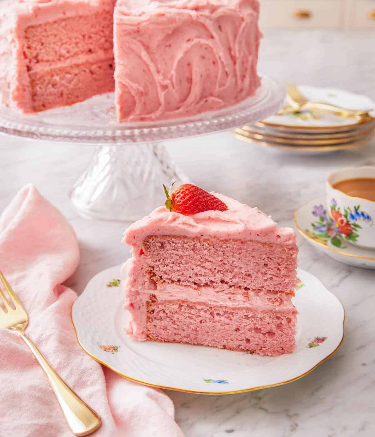 A slice of strawberry cake on a plate with a fresh strawberry on top with a cake stand with the rest of the cake in the back.