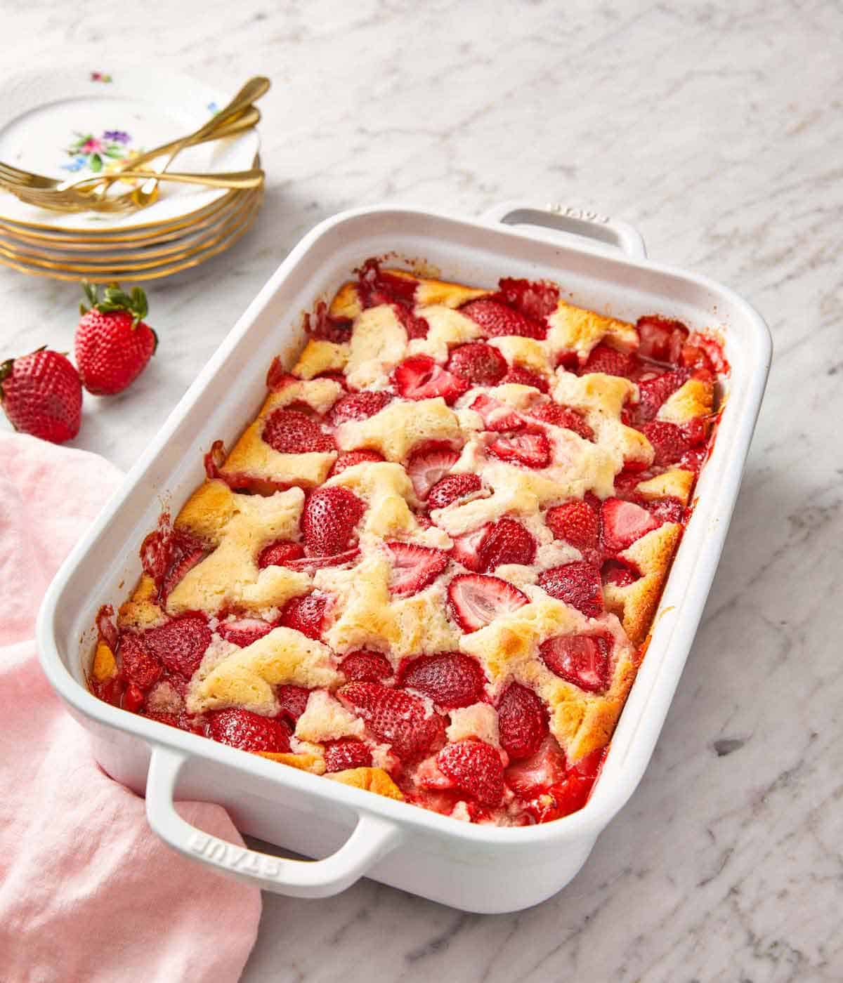 A white baking dish of strawberry cobbler with a stack of plates and forks in the background.