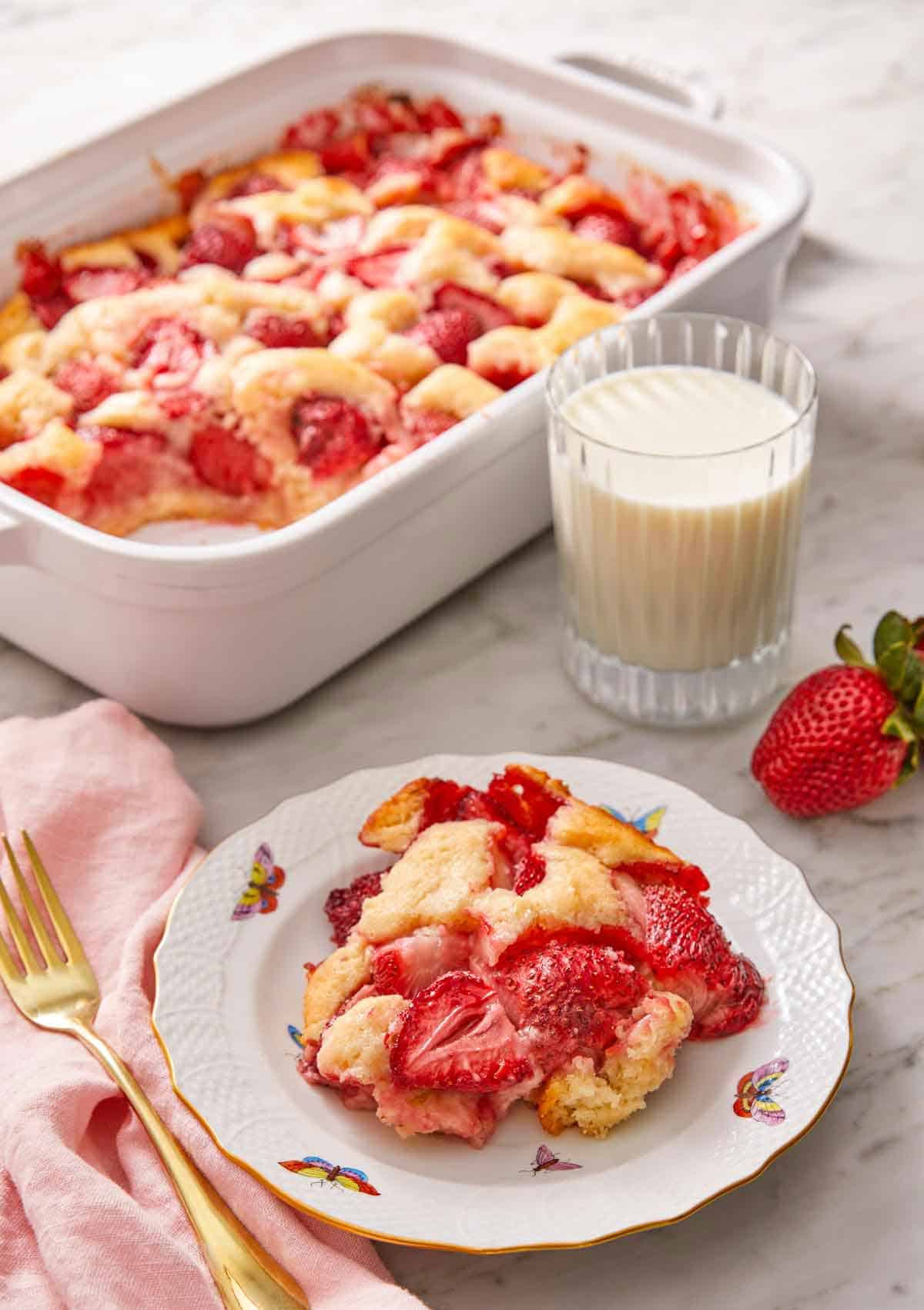 A plate with a serving of strawberry cobbler in front of a glass of milk and baking dish.