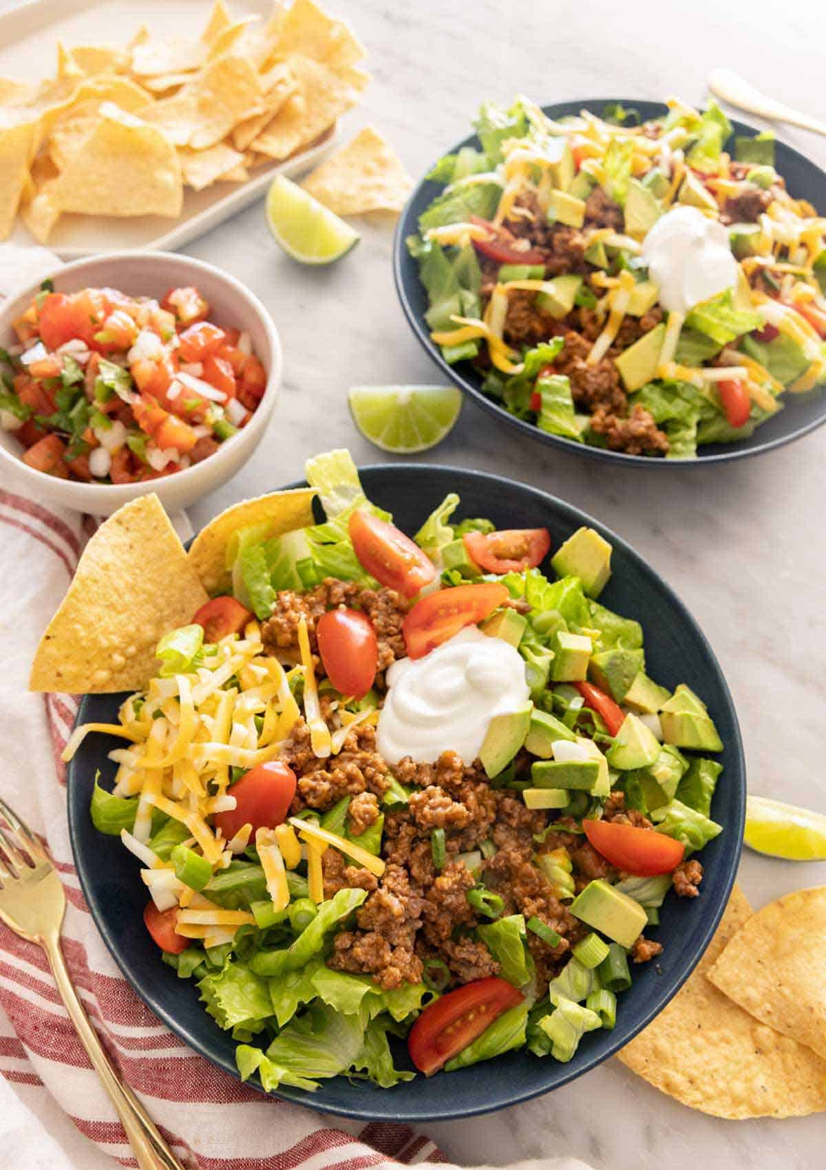 Two bowls of taco salad with a small bowl of pico de gallo and chips on the side.
