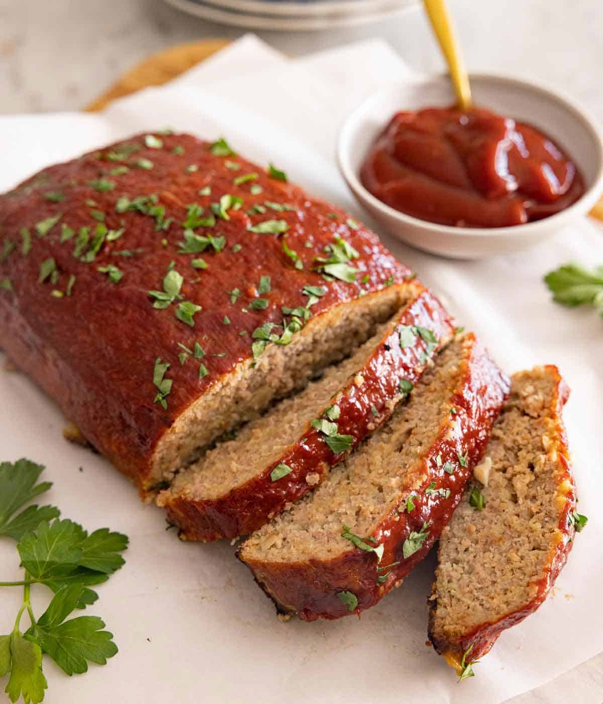 A loaf of turkey meatloaf with three slices cut with a bowl of sauce in the background.