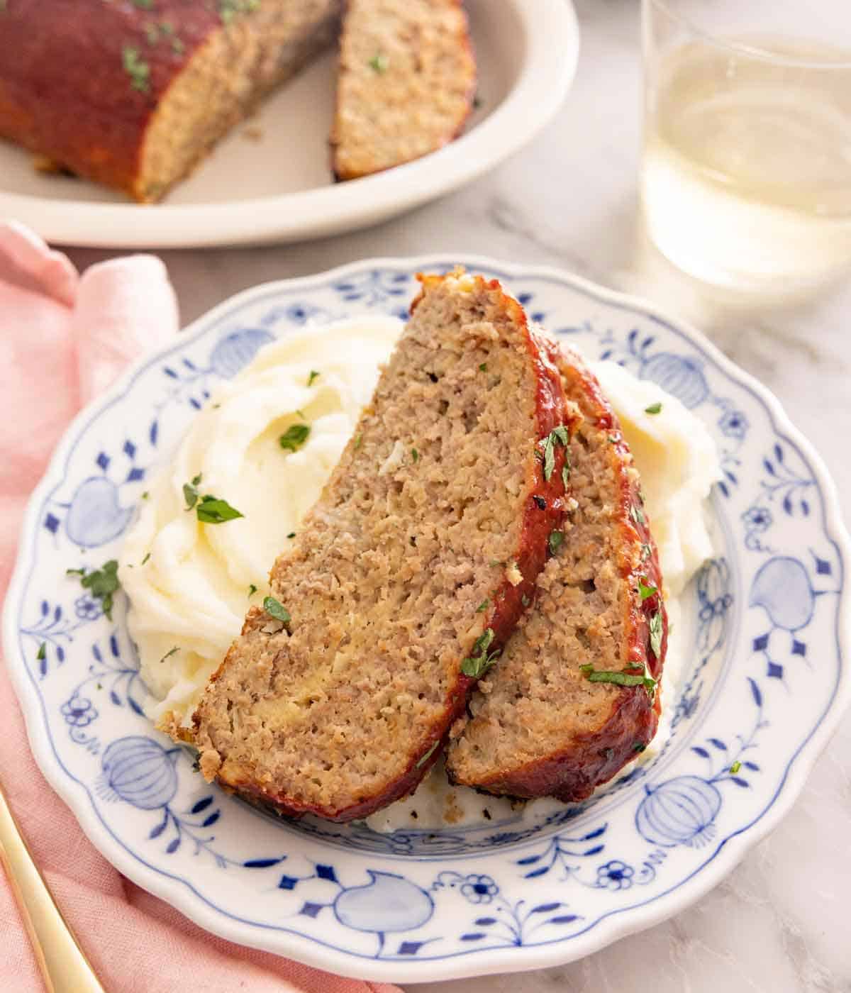 A plate of mashed potatoes with two slices of turkey meatloaf placed on top with some chopped herb garnish.