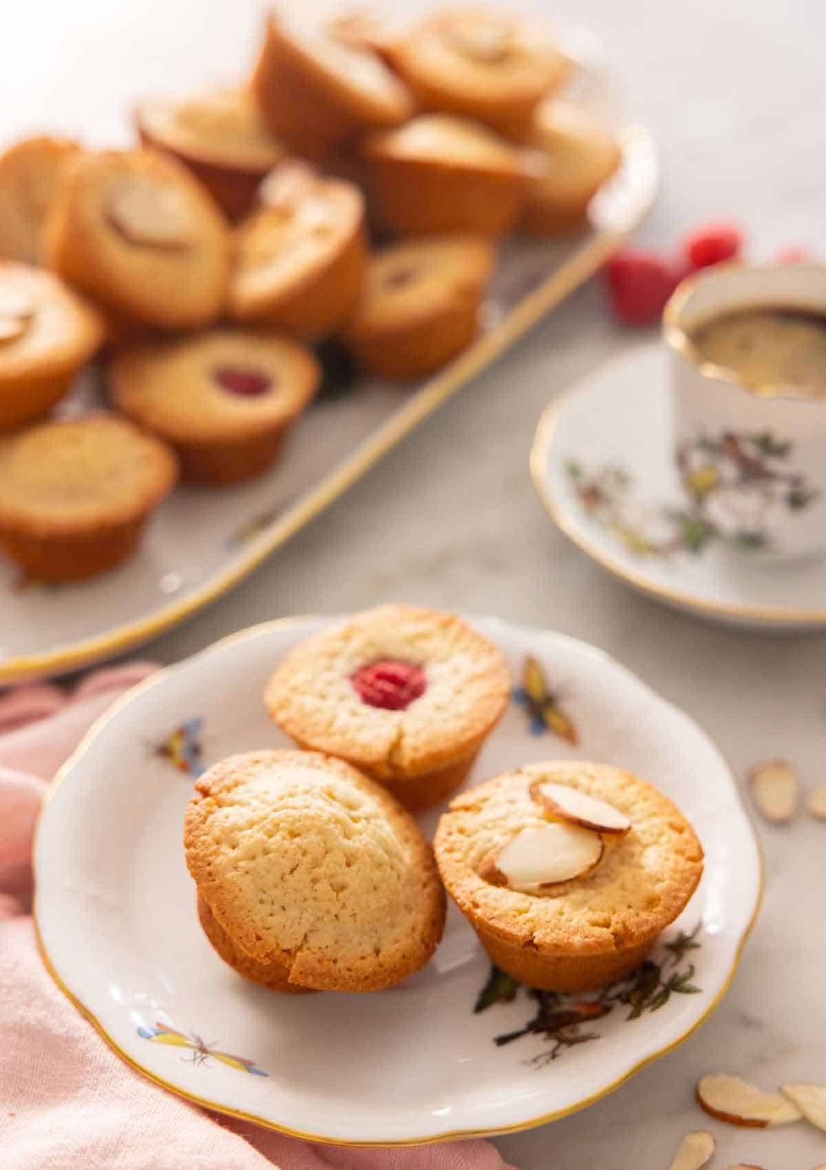 A plate with three financiers on a plate, one plain, one with a raspberry on top, and one with almonds on top.